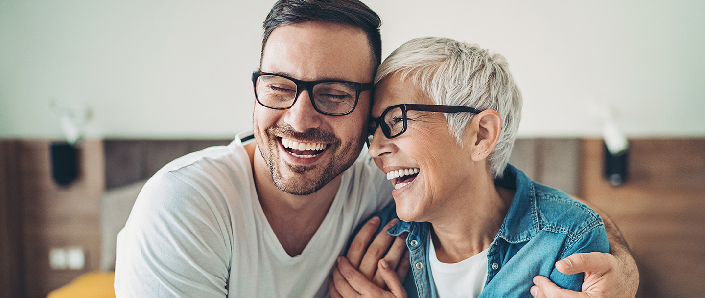 Adult mother and son, sitting in the living room sharing a laugh. The son has his arms around his mother, their heads leaned in.