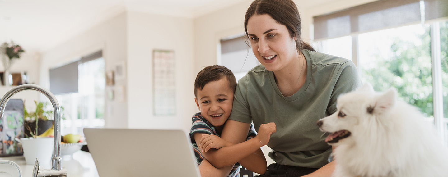Woman at her kitchen counter looking information up on her laptop with her son hugging her arm and a fluffy, white dog looking on